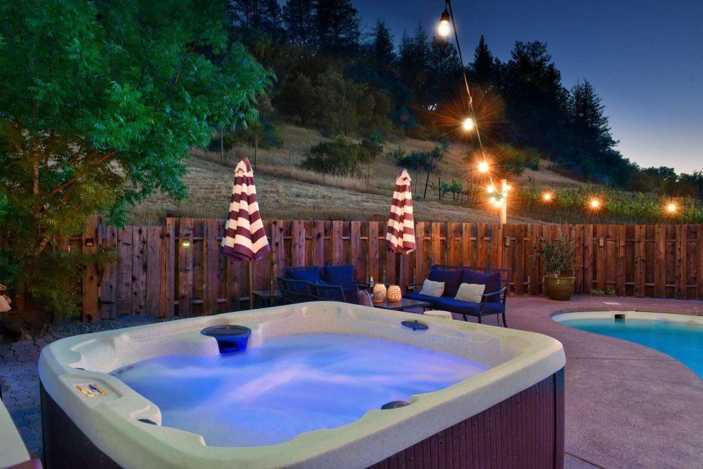 A hot tub with blue LED lighting sits next to a private pool in a fenced backyard