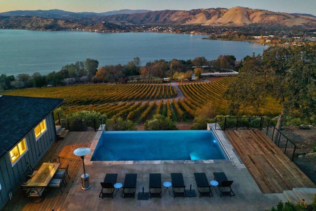A modern lakefront patio with a private pool overlooks a vineyard, with a large lake and rolling hills in the background at sunset. Lounge chairs and a dining area offer relaxation in one of the finest Northern California vacation rentals.
