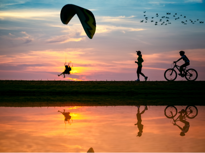 Silhouettes of a paraglider, a runner, and a cyclist on a hill at sunset, with colorful sky, flying birds, and their reflections in a calm body of water below.